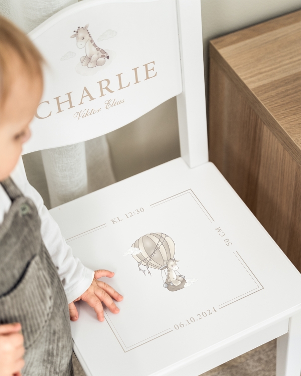 Hvit dåpsstol med navn på ryggstøtten, samt dato, klokkeslett og sjiraffmotiv på setet